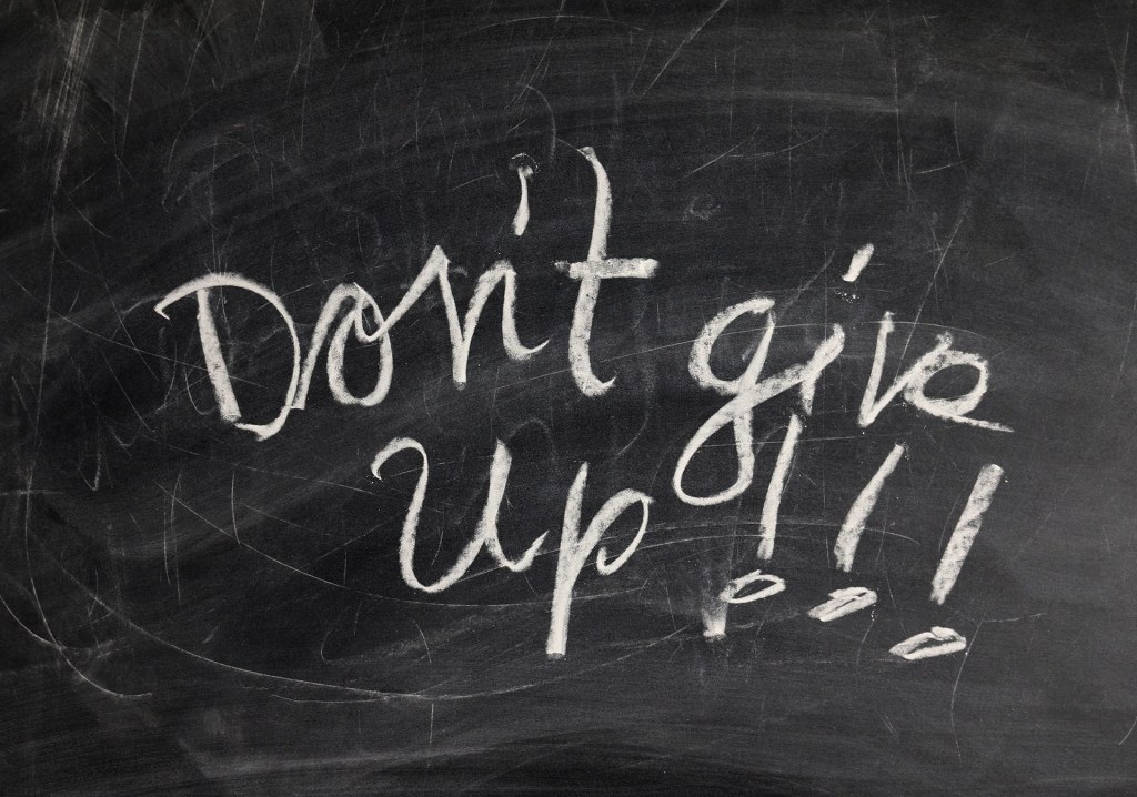 Image of a well used chalkboard with the words "Don't give up!!!" written on it in white chalk, in script lettering.