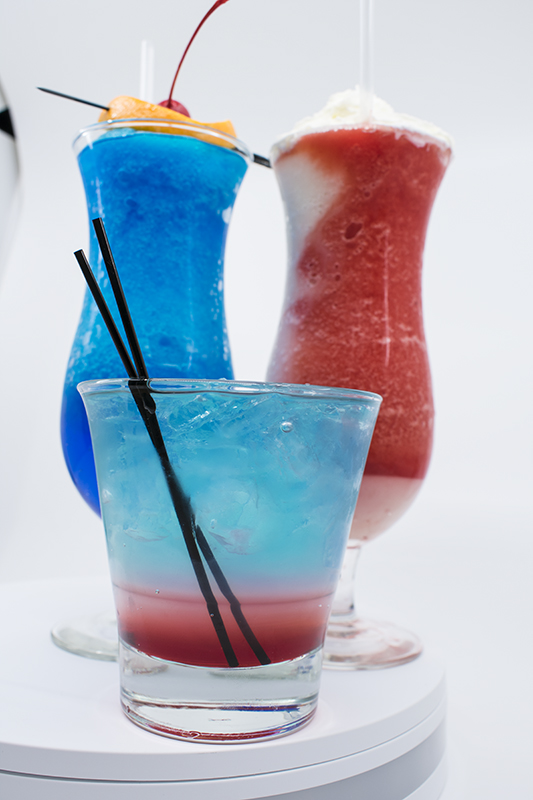Photo of a trio of cocktails taken in a lightbox. The two cocktails in the background are in tall dacquiri glasses; one blue and one red and white. The cocktail in the foreground is in a rocks glass, blue at the top and shading down into pink and red.